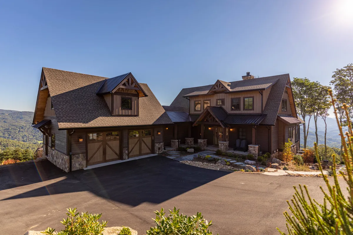 The image showcases the main entrance of Almost Heaven at Eagles Nest, featuring a welcoming front porch seating area, striking stone detailing on the facade, and a beautifully landscaped garden. The mountain backdrop is subtly visible, evoking the natural setting of the resort.