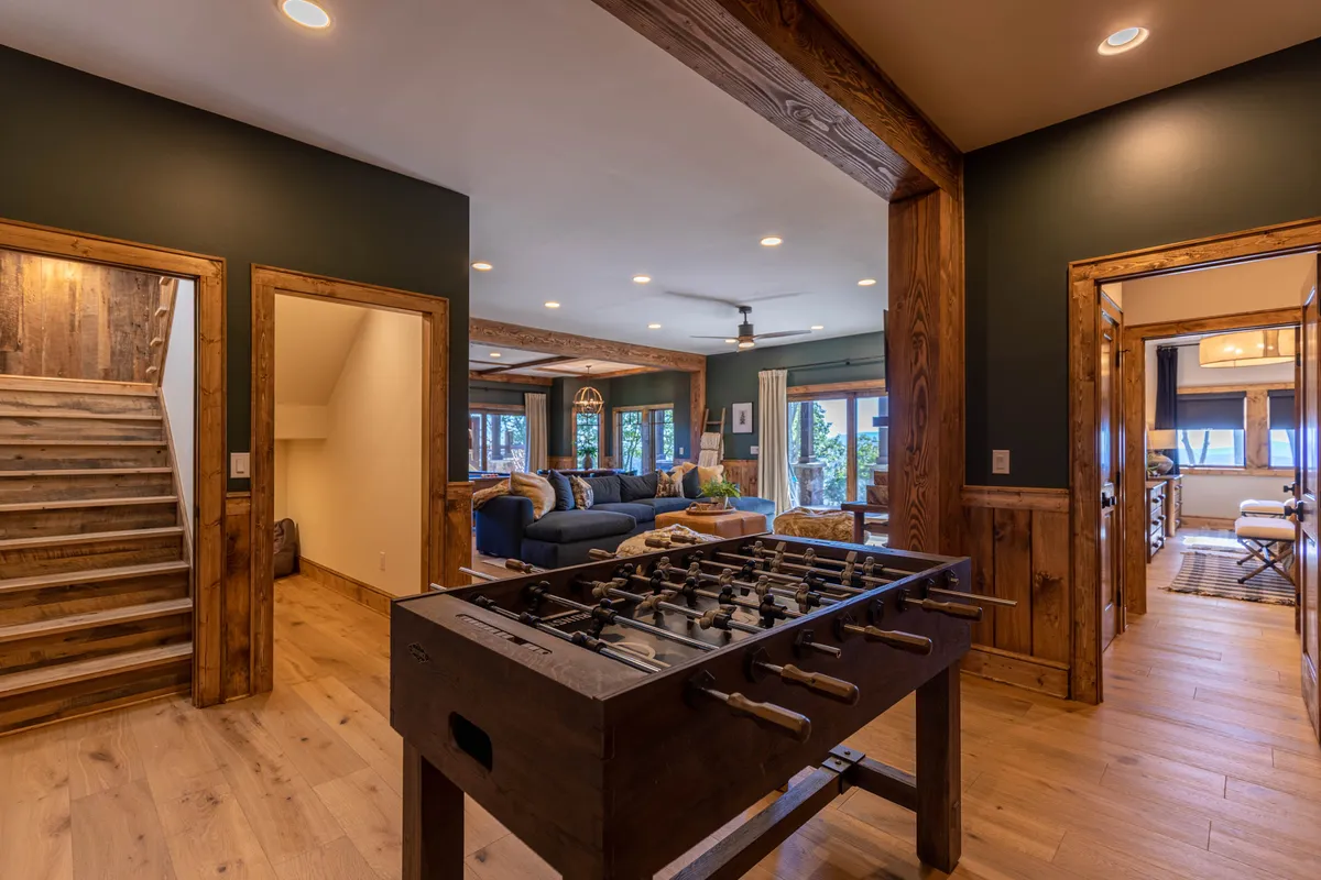 This image showcases a lively and sunlit game room featuring a foosball table, a cozy blue couch, and large windows that let in natural light. The wooden staircase and open layout suggest ample space for social gatherings and family activities, making it the hub for fun and friendly competition.