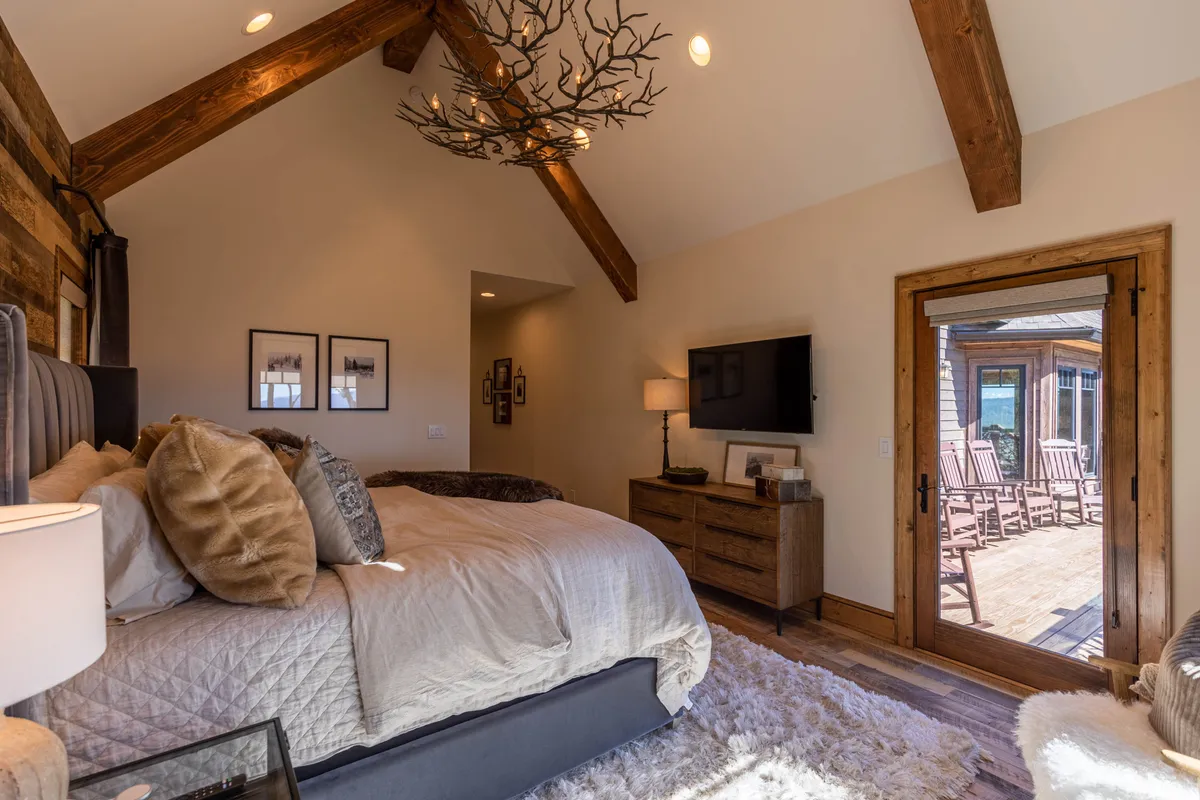 A tranquil bedroom showcasing a large, soft bed with luxurious bedding, complemented by an antler chandelier and a wooden dresser. A glass sliding door opens to the deck, inviting natural light and access to the outdoors, while the room balances coziness with a sense of openness.