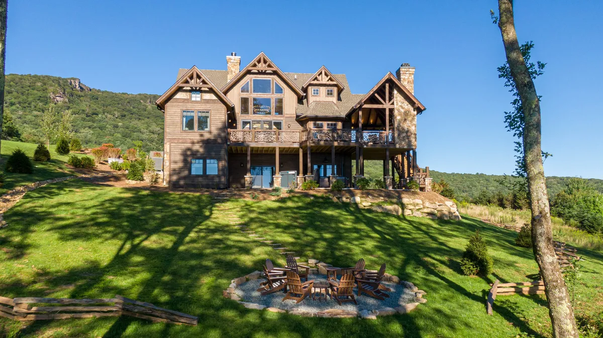 A wide-angle view of the large wooden house set against a mountain backdrop, with a spacious outdoor seating area surrounded by greenery. The setting invites family gatherings, outdoor barbecues, and time spent enjoying nature.
