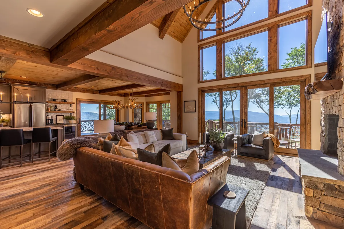 A spacious living area featuring large windows that frame breathtaking mountain views, a leather sofa arranged around a stone fireplace, and rustic wooden beams overhead. A chandelier adds a touch of elegance to the room, which is designed for both relaxation and gatherings.