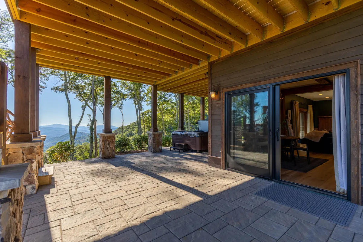 A spacious patio with a hot tub overlooking expansive mountain views. The patio features comfortable seating and space for social gatherings, with a sliding glass door connecting the outdoor and indoor living areas. The serene atmosphere is perfect for relaxing, enjoying wine, or evening conversations with friends against a beautiful natural backdrop.