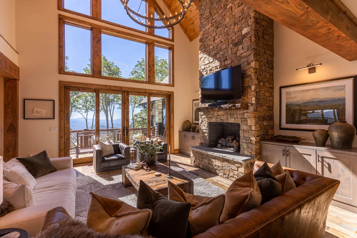 A warm and inviting living room with a large stone fireplace as the focal point, expansive windows letting in natural light and views, a rustic wooden coffee table, and comfortable seating arranged for group conversation or lounging.