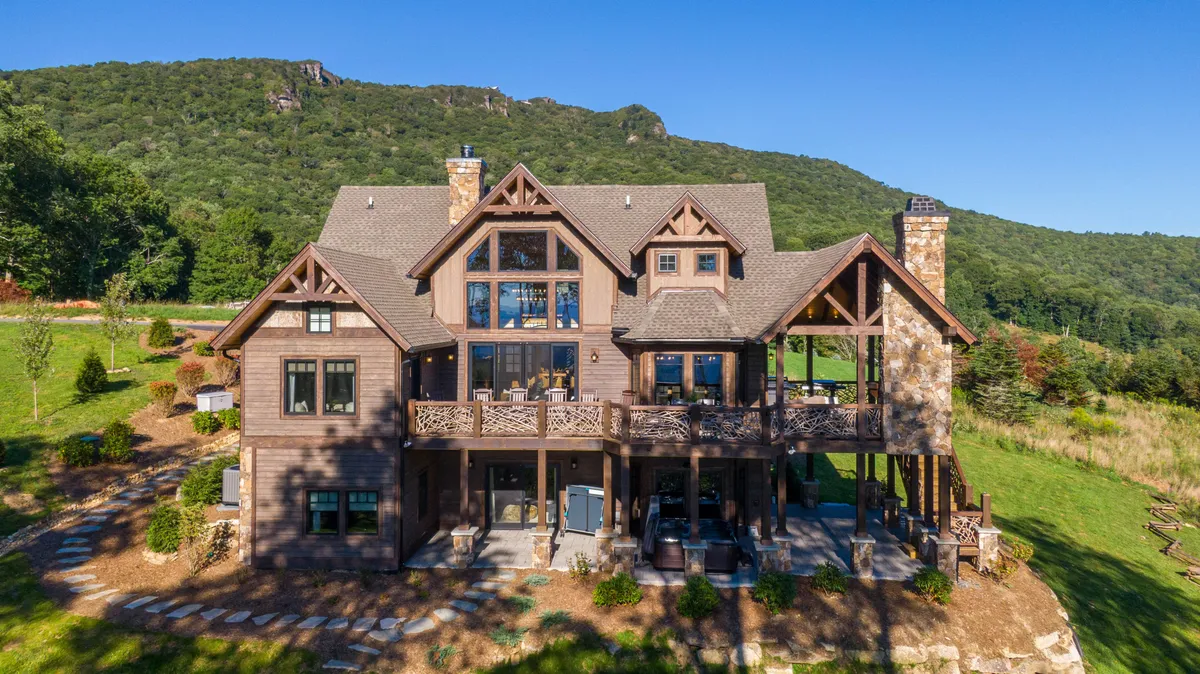 The image showcases a luxurious mountain home with a spacious wooden deck, dramatic stone chimney, and expansive windows overlooking panoramic Blue Ridge Mountain views. Lush greenery surrounds the property, and the deck is perfectly positioned for outdoor dining, sunset watching, and family gatherings in nature.